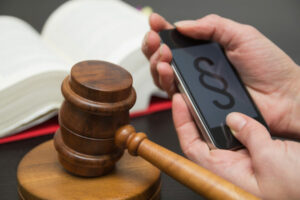 Richterhammer, Gesetzbuch und zwei Händer halten ein Smartphone. Darauf ist ein Paragraph zu sehen.